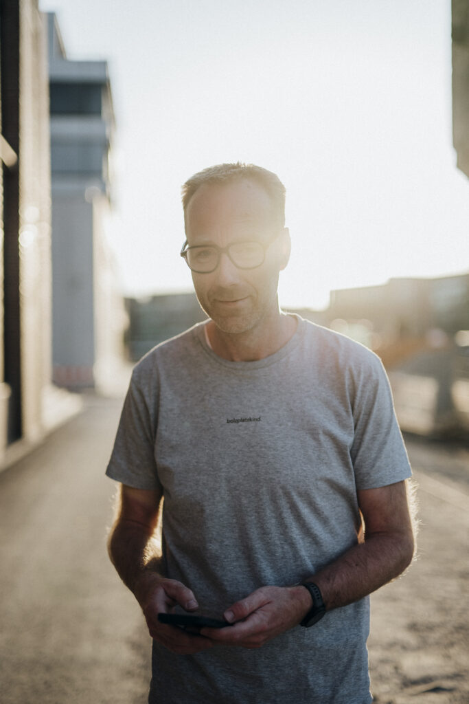 Mann mit Brille in grauem T-Shirt hält Smartphone, Sonnenuntergang im Hintergrund.