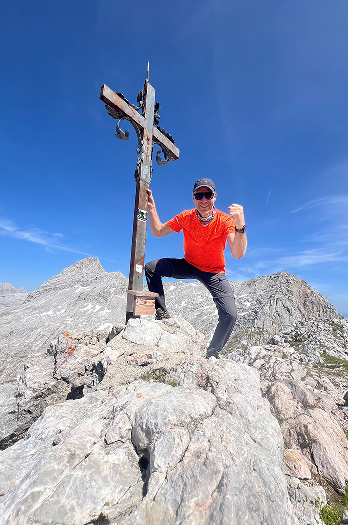Eine Person in orangefarbenem Hemd lächelt erfreut auf einem felsigen Berggipfel neben einem Holzkreuz.