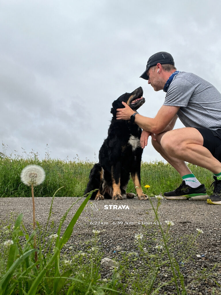Laechelnder Mann in Grau und Schwarz kniet bei schwarzem Hund auf Kiesweg