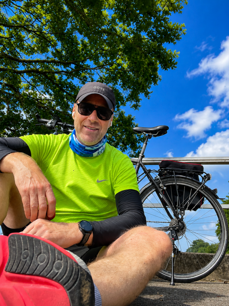 Radfahrer laechelt entspannt in neongruen neben schwarzem Rad und blauem Himmel