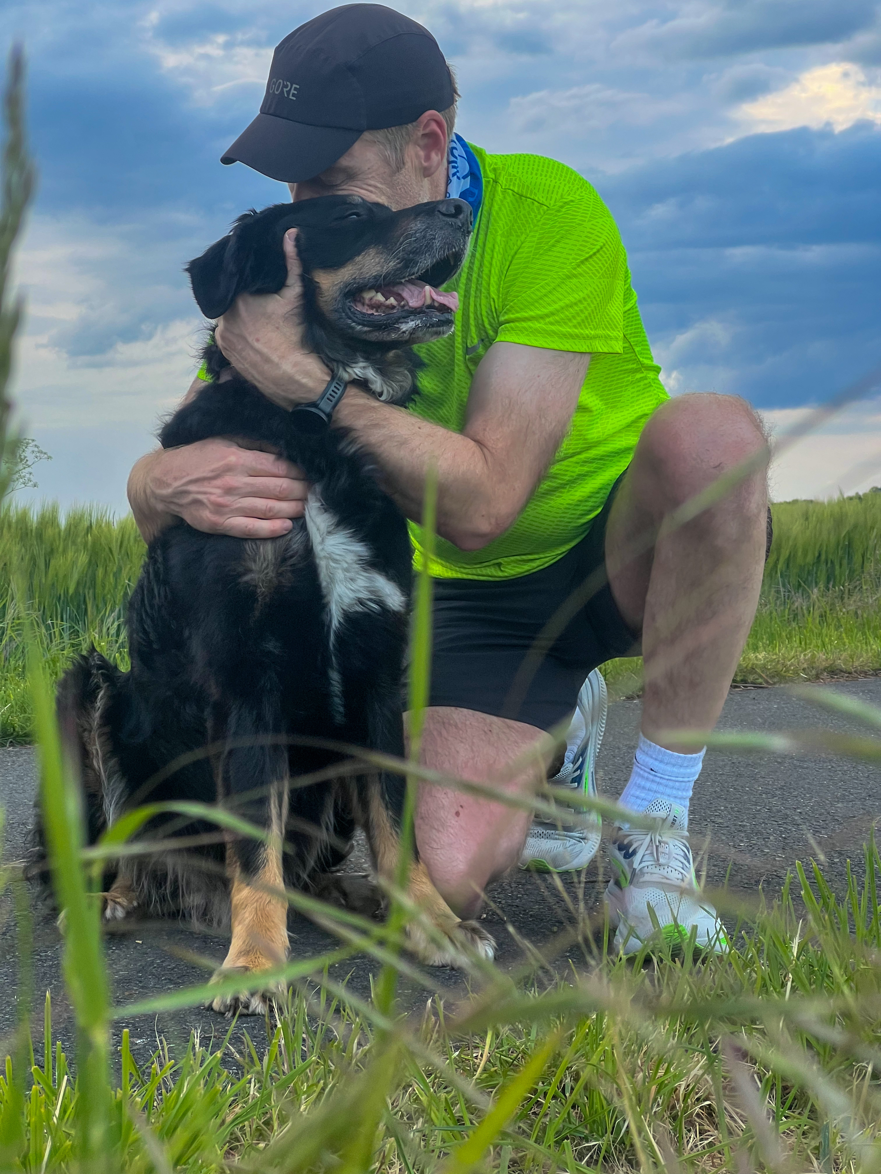 Laechelnder Mann in neon Gruen umarmt schwarzen Hund mit weisser Brust im Gras