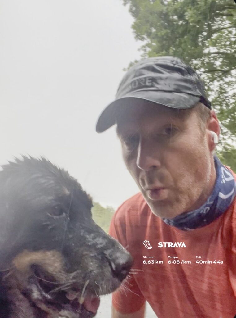 Mann mit neutralem Blick in rotem Shirt und Kappe neben schwarzem Hund im Nebel