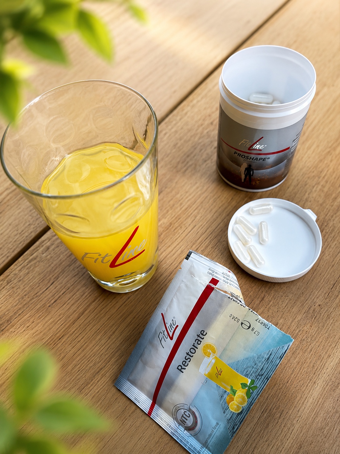 FitLine Glas mit orangefarbenem Getraenk neben Kapseln und Restorate Beutel